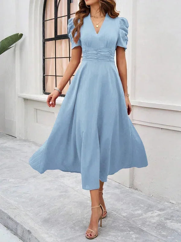 Woman wearing a light blue dress standing in front of a white wall with a window.
