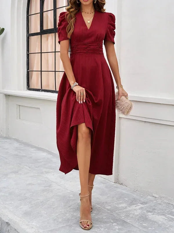 Woman wearing a burgundy dress with puff sleeves and a high-low hem, standing against a white wall.