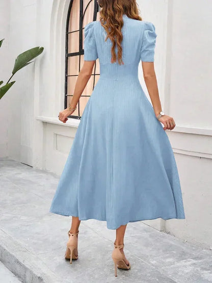 Woman wearing a light blue dress standing in a room with white walls and a window.