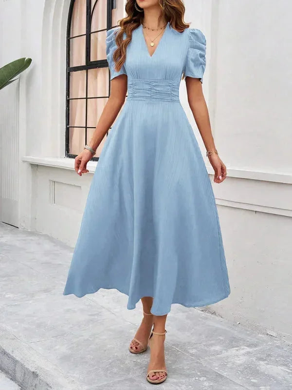 Woman wearing a light blue dress standing in front of a white wall with a window.