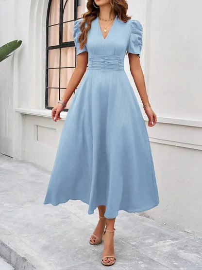 Woman wearing a light blue dress standing in front of a white wall with a window.