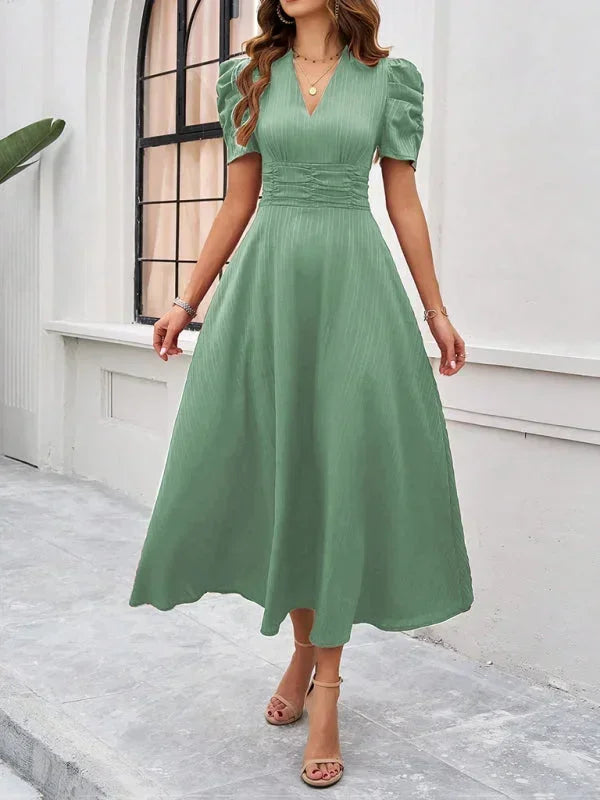 Woman wearing a green dress standing in front of a white wall with a window.