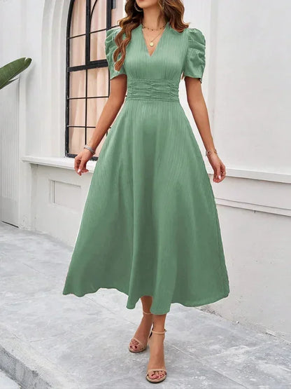 Woman wearing a green dress standing in front of a white wall with a window.