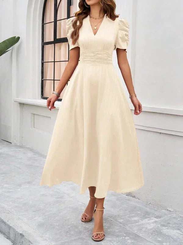 Woman wearing a beige dress standing in front of a white wall.