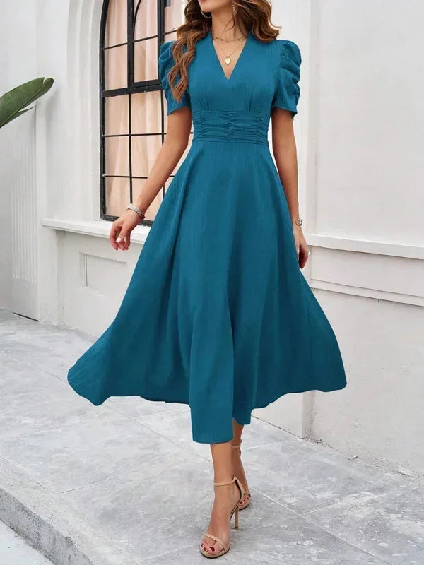Woman wearing a teal dress standing in front of a white wall.
