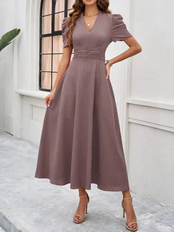 Woman wearing a long, light brown dress standing against a white wall.