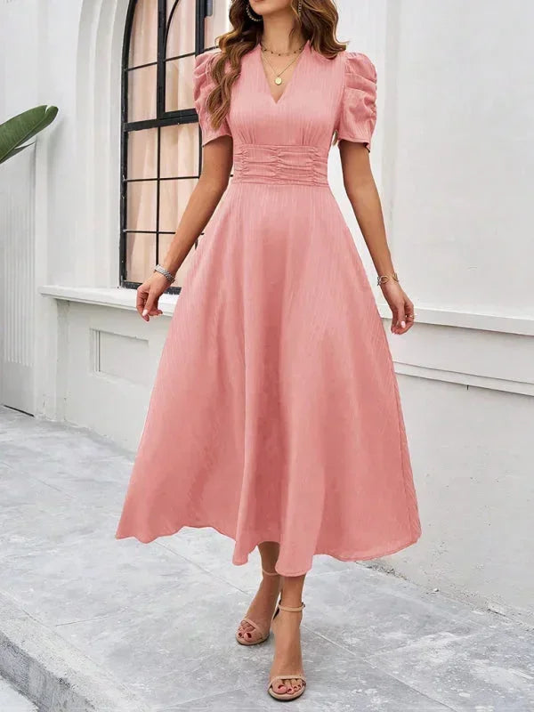 Woman wearing a pink dress standing on a stone path with a white wall and window in the background.