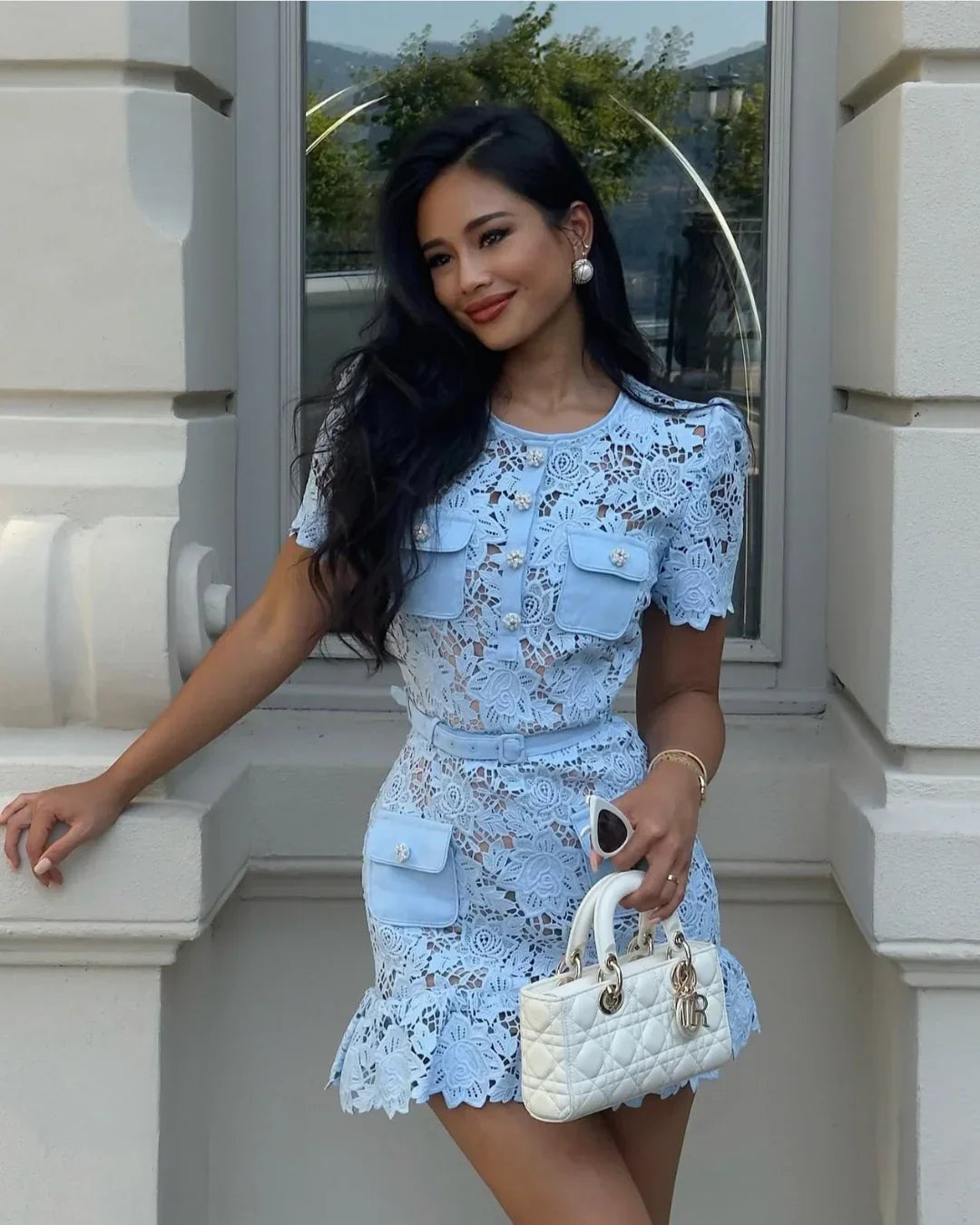 Woman in a light blue lace dress holding a white handbag against a building background