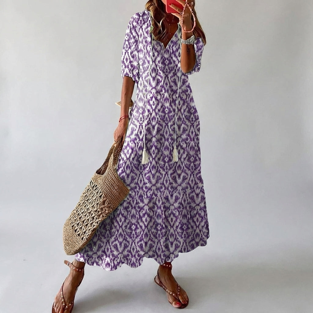 Woman wearing a purple patterned dress holding a woven bag against a plain background